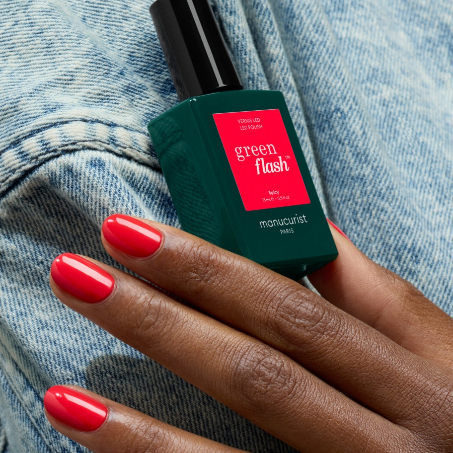 A hand with bright red-painted nails holds a bottle of Spicy by Manucurist, evoking an eighties nail polish vibe against a background of light blue denim fabric.
