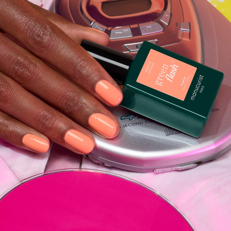 A hand with Tangerine polish by Manucurist rests on a portable nineties CD player, with a bottle of Tangerine nearby and a pink CD partially visible in the foreground.