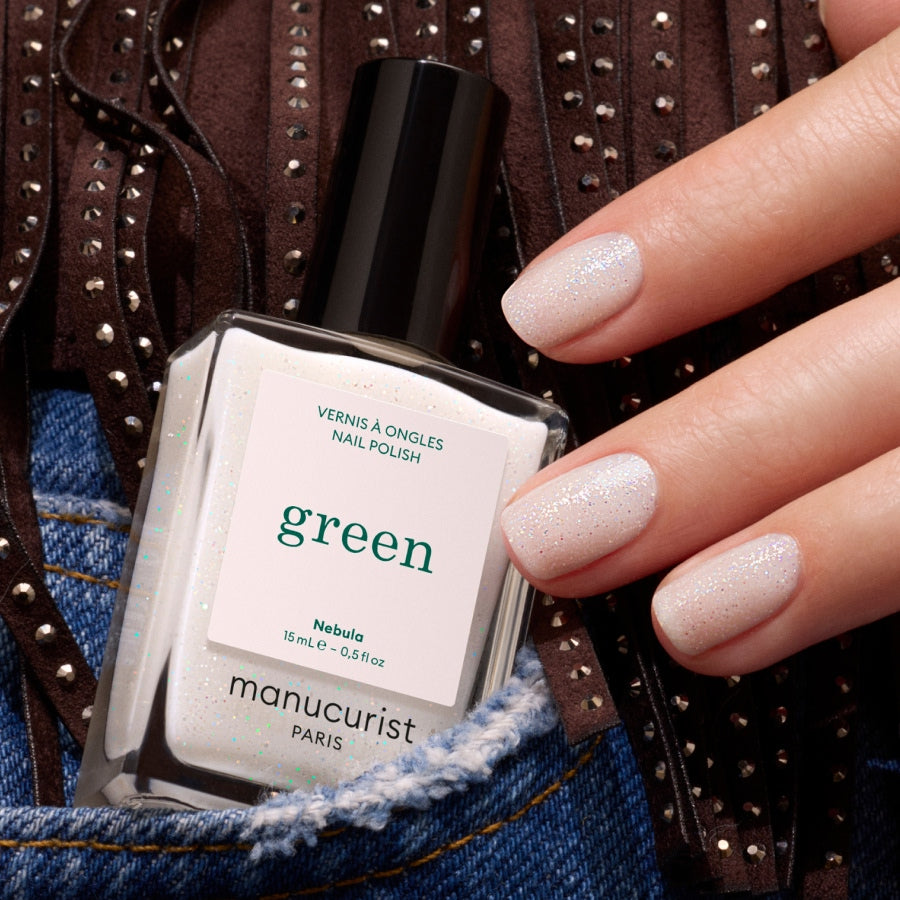 A hand with light, shimmery holographic nail polish holds a bottle of Manucurist Nebula, set against blue denim and brown fringed fabric.