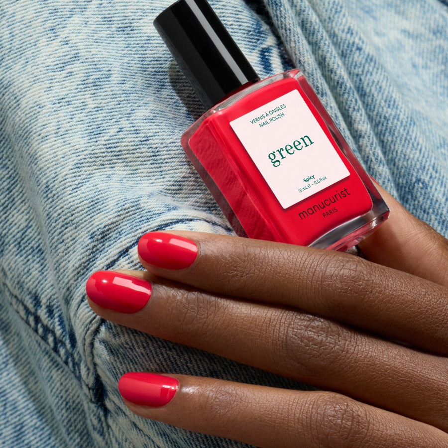A hand with neatly manicured nails painted in Spicy by Manucurist holds the bottle against a light blue denim background.