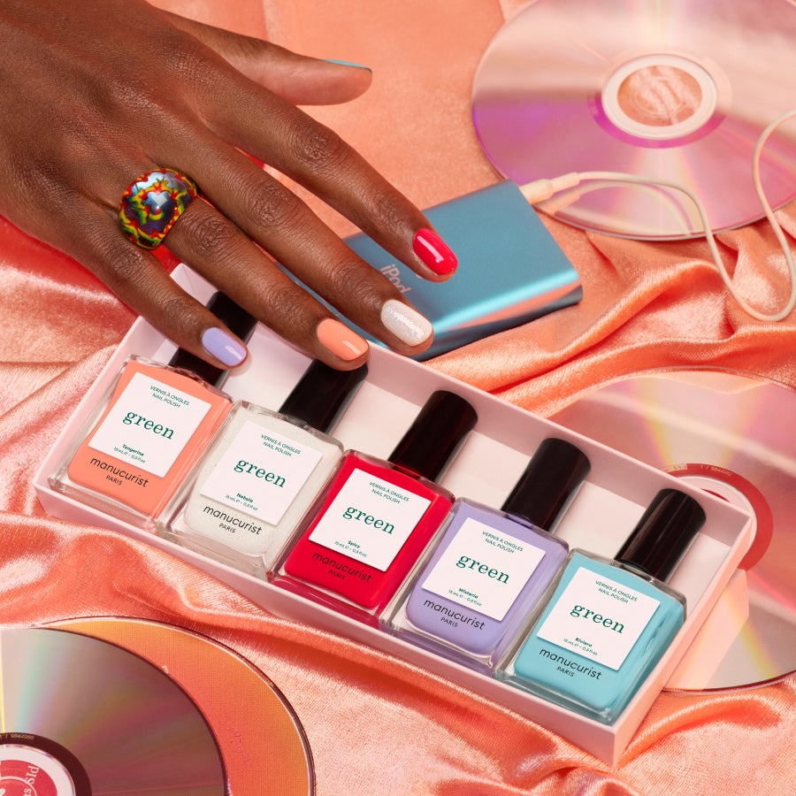 A hand with multicolored nails and a beaded ring rests on satin beside CDs. In front, Manucurist Rainbow Collections—five nail polishes in curated palettes—are displayed in a sleek white box.