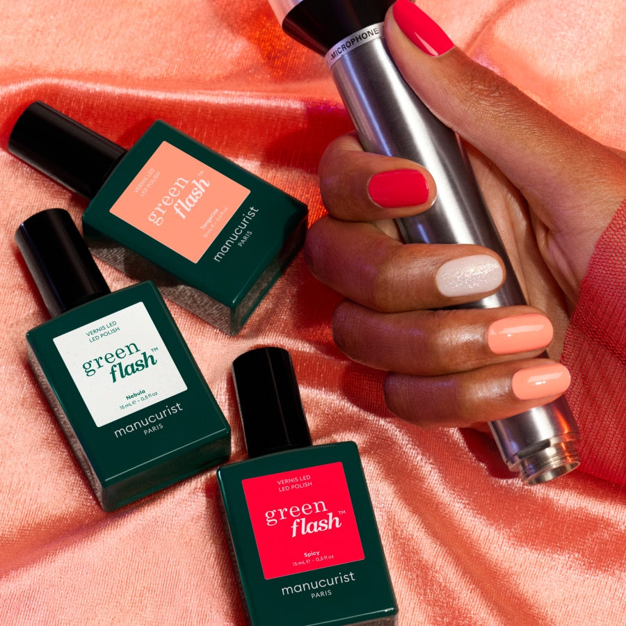 A hand with pink, red, and nude manicured nails holds a metallic nail lamp. Three Manucurist Trio Collections bottles rest on pink satin, highlighting curated colours inspired by the latest nail trends.