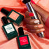 A hand with pink, red, and nude manicured nails holds a metallic nail lamp. Three Manucurist Trio Collections bottles rest on pink satin, highlighting curated colours inspired by the latest nail trends.