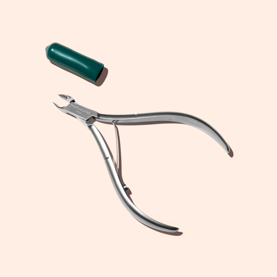 A pair of Manucurist Cuticle Nippers, crafted from professional-quality stainless steel, is shown with a green plastic cap nearby on a light beige background.