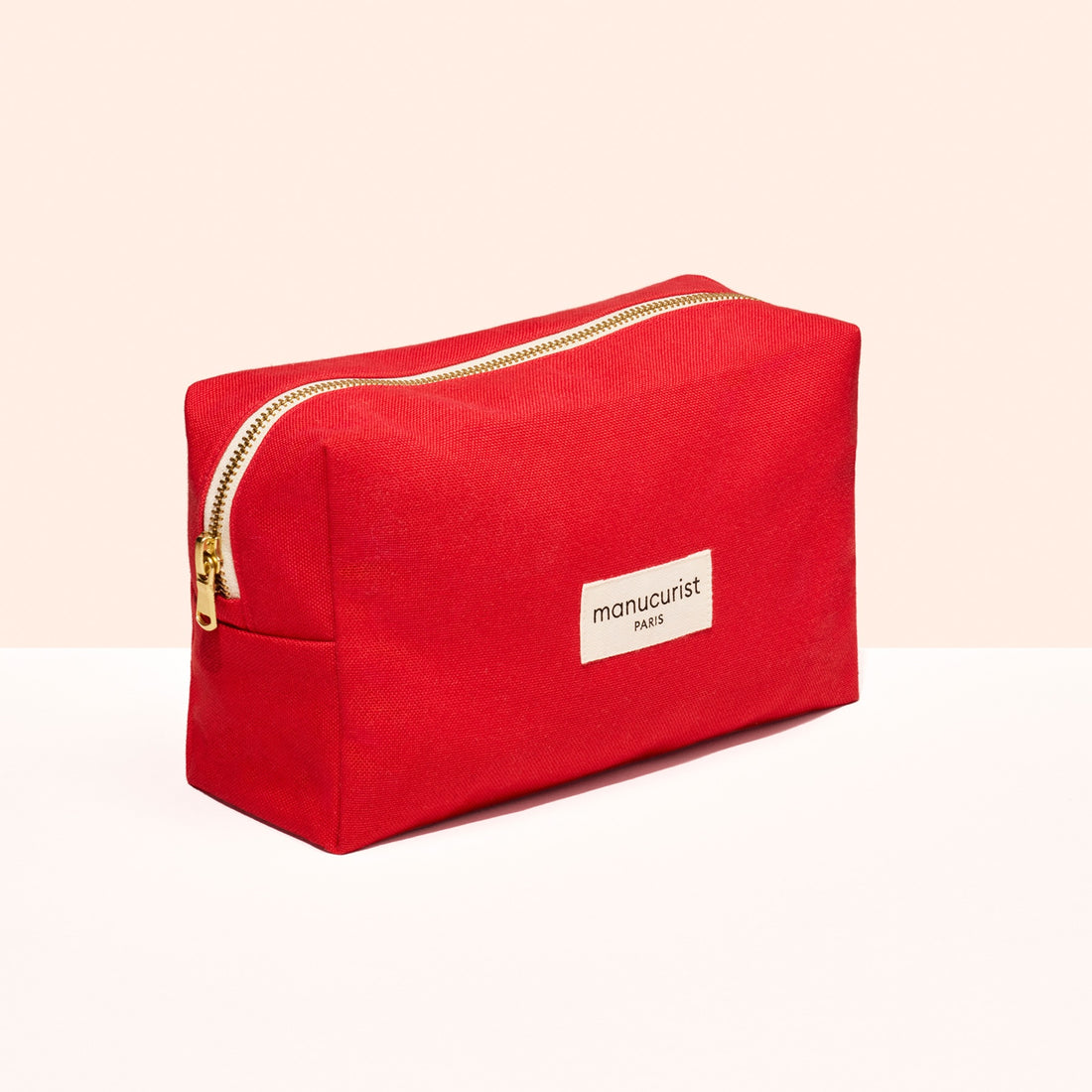 The Red Organic Cotton Pouch by Manucurist features a bright red rectangular shape, gold zipper, and white "manucurist PARIS" label, shown on a white and light peach background.
