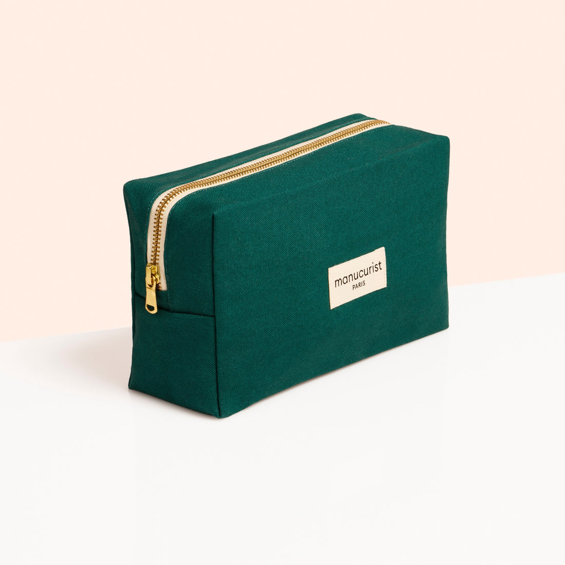 A green rectangular Manucurist organic cotton pouch with a gold zipper and white label stands on a white surface against a soft peach background.