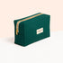 A green rectangular Manucurist organic cotton pouch with a gold zipper and white label stands on a white surface against a soft peach background.