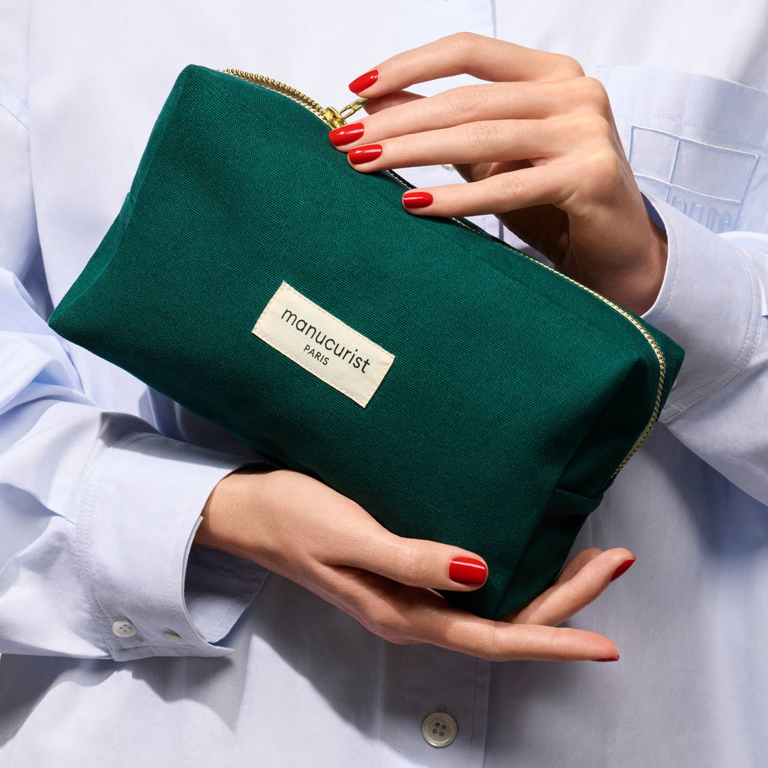 A person with red nails holds a Manucurist Organic cotton pouch, wearing a light blue button-up shirt.