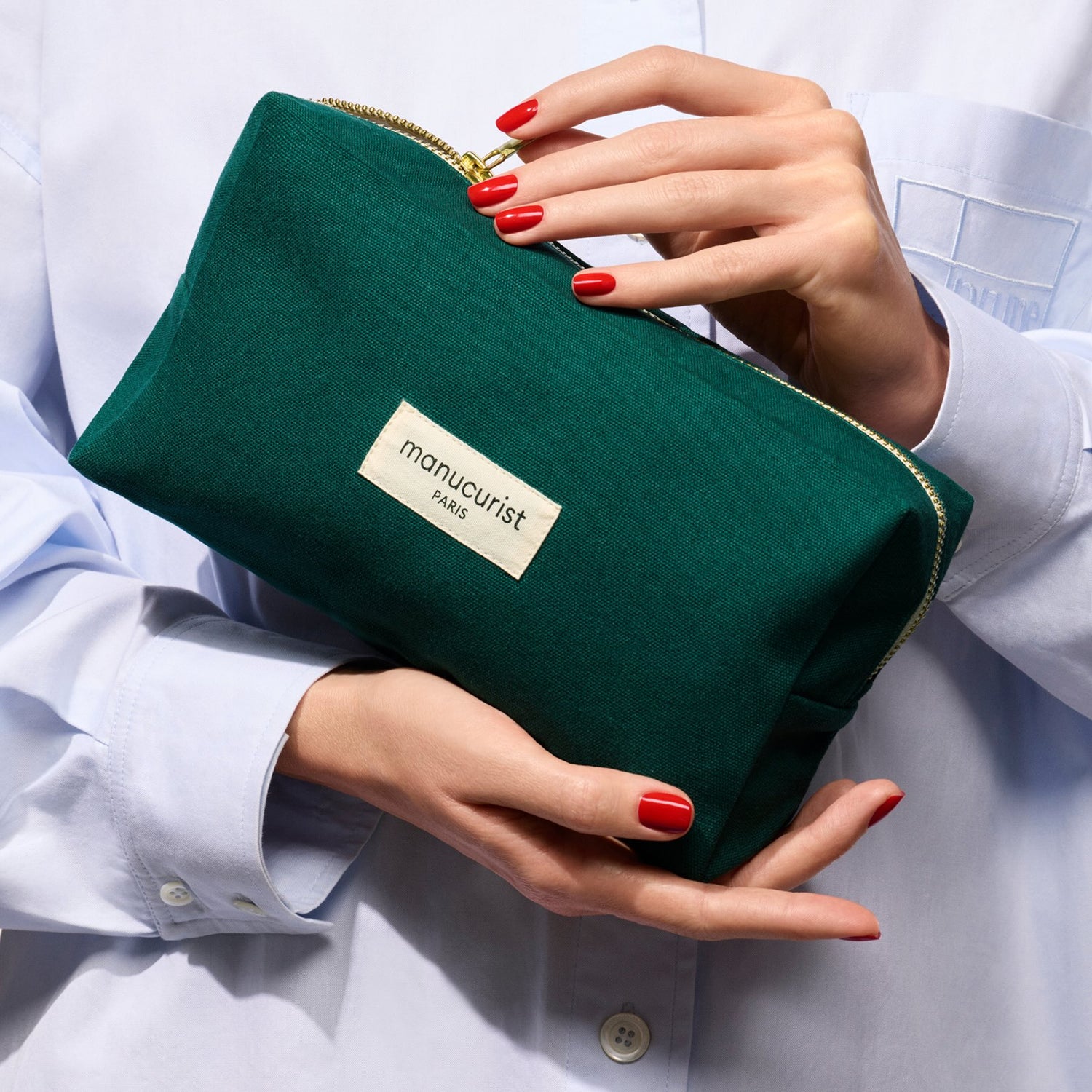 A person with red nails holds a Manucurist Organic cotton pouch, wearing a light blue button-up shirt.