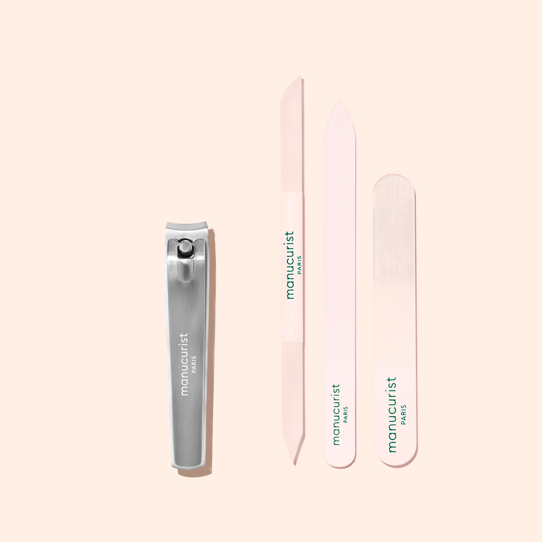 A Manucurist Nail Clipper in silver and three light pink nail files are arranged vertically on a pale pink background, each labeled “Manucurist Paris” in green text—perfect for achieving flawless natural nails.