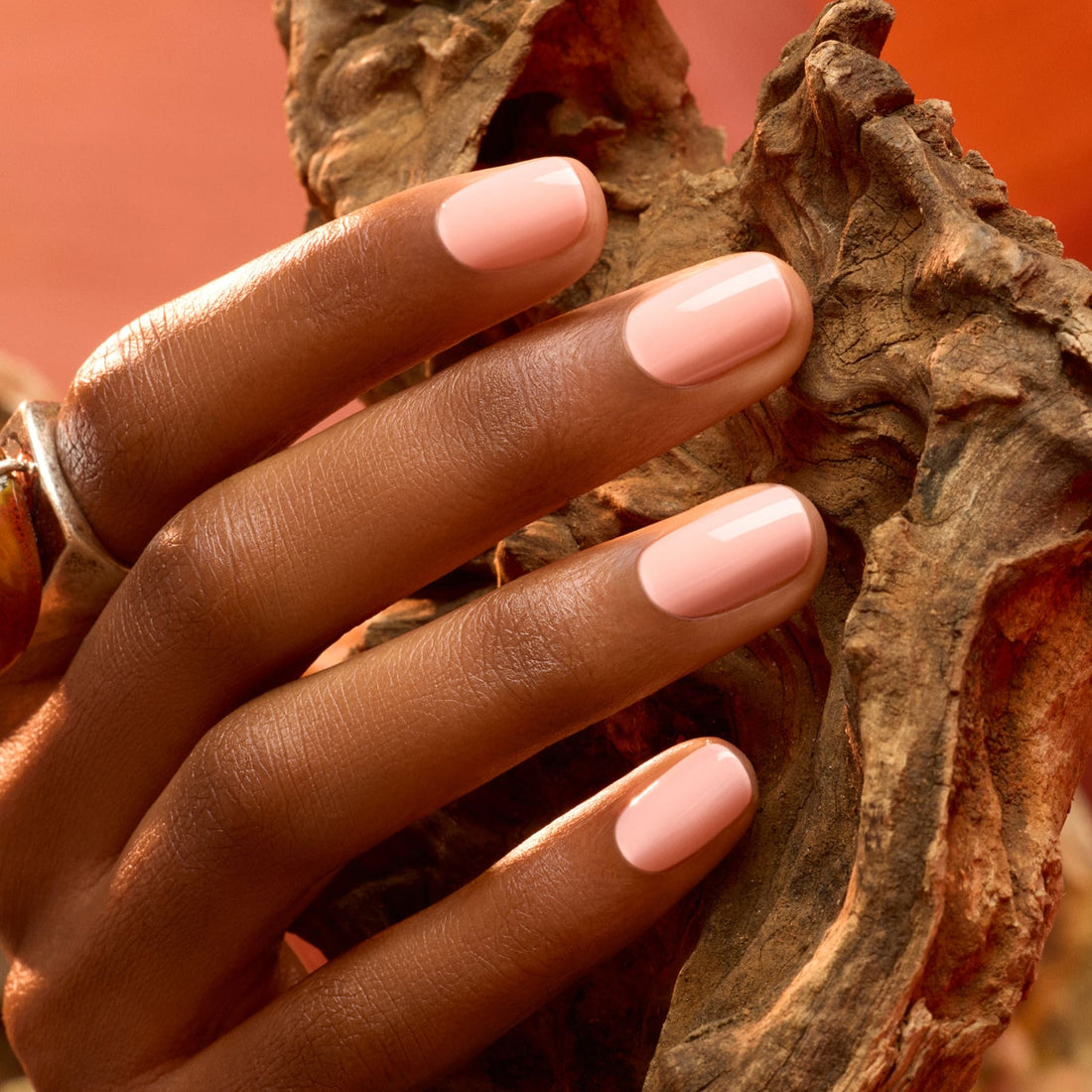 A hand with neatly manicured nails painted in Manucurist’s Milky Peach polish rests against textured brown wood, warm lighting highlighting the smooth skin and the glossy finish of the manicure.