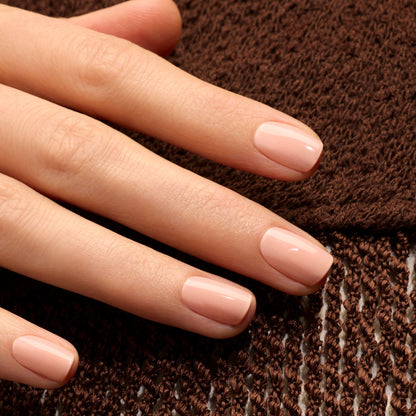 A hand with short, neatly manicured nails painted in Manucurist’s Milky Peach, a glossy orange beige shade, rests on textured brown fabric.