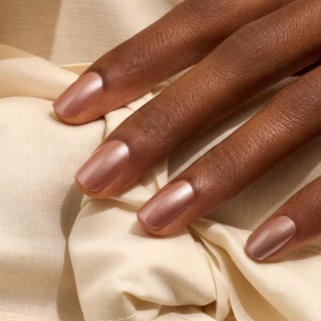 A hand with neatly manicured nails painted in Manucurist’s Sandstone, a pearly brown nail polish, rests on a soft, cream-colored fabric.