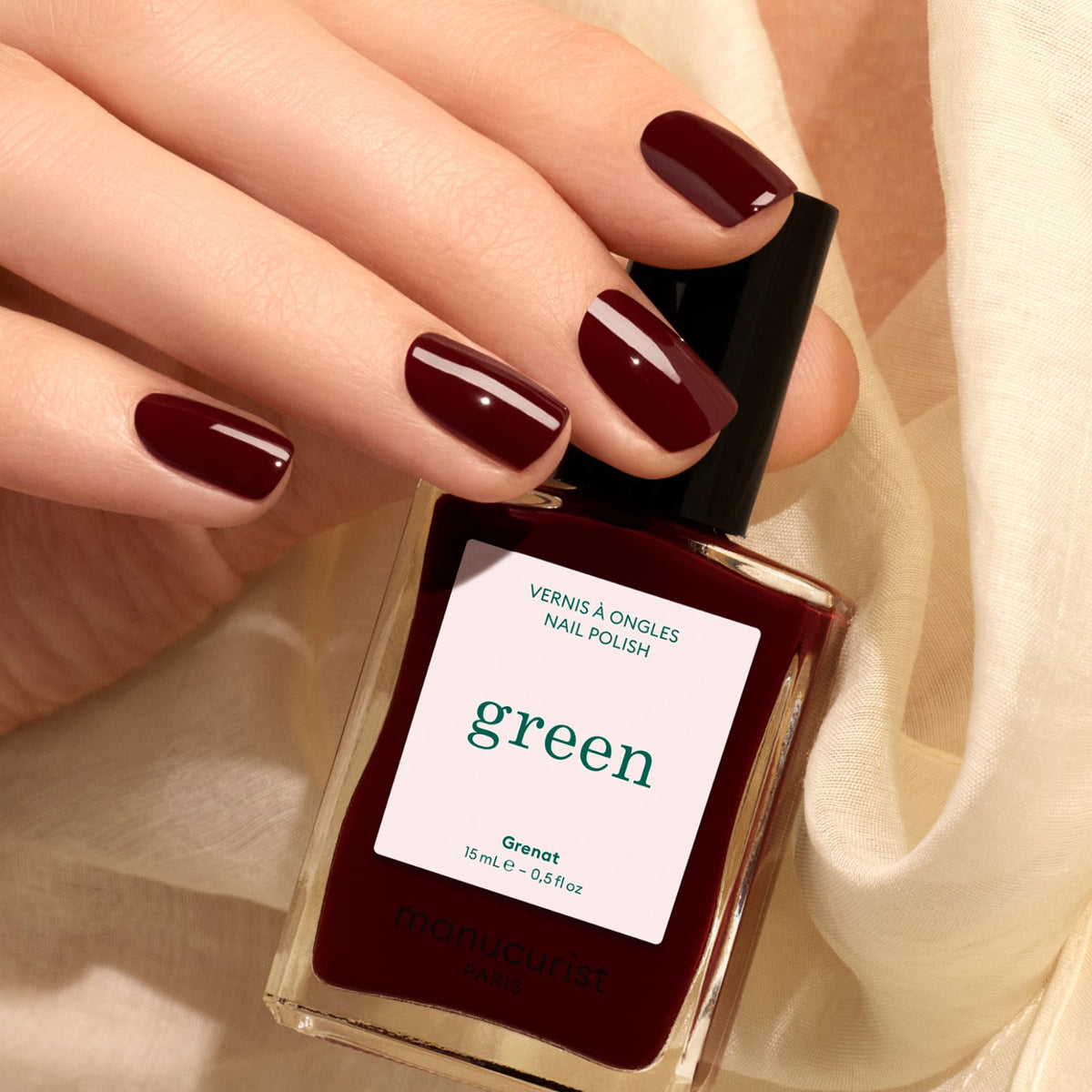 A hand with burgundy painted nails holds a matching bottle of Manucurist Grenat nail polish. Cream-colored fabric is in the background.