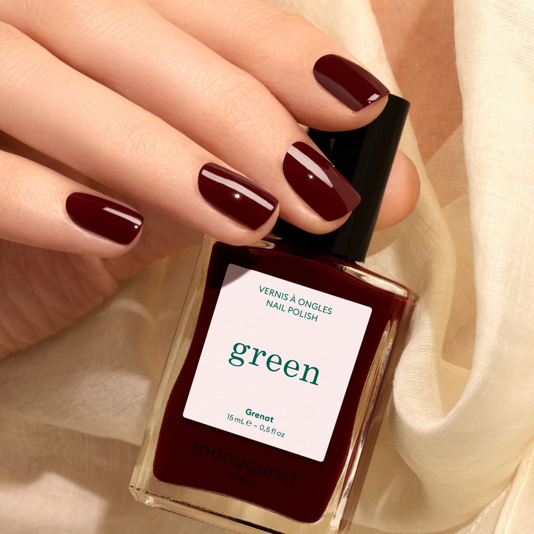 A hand with burgundy painted nails holds a matching bottle of Manucurist Grenat nail polish. Cream-colored fabric is in the background.