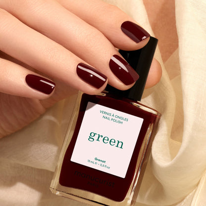 A hand with burgundy painted nails holds a matching bottle of Manucurist Grenat nail polish. Cream-colored fabric is in the background.