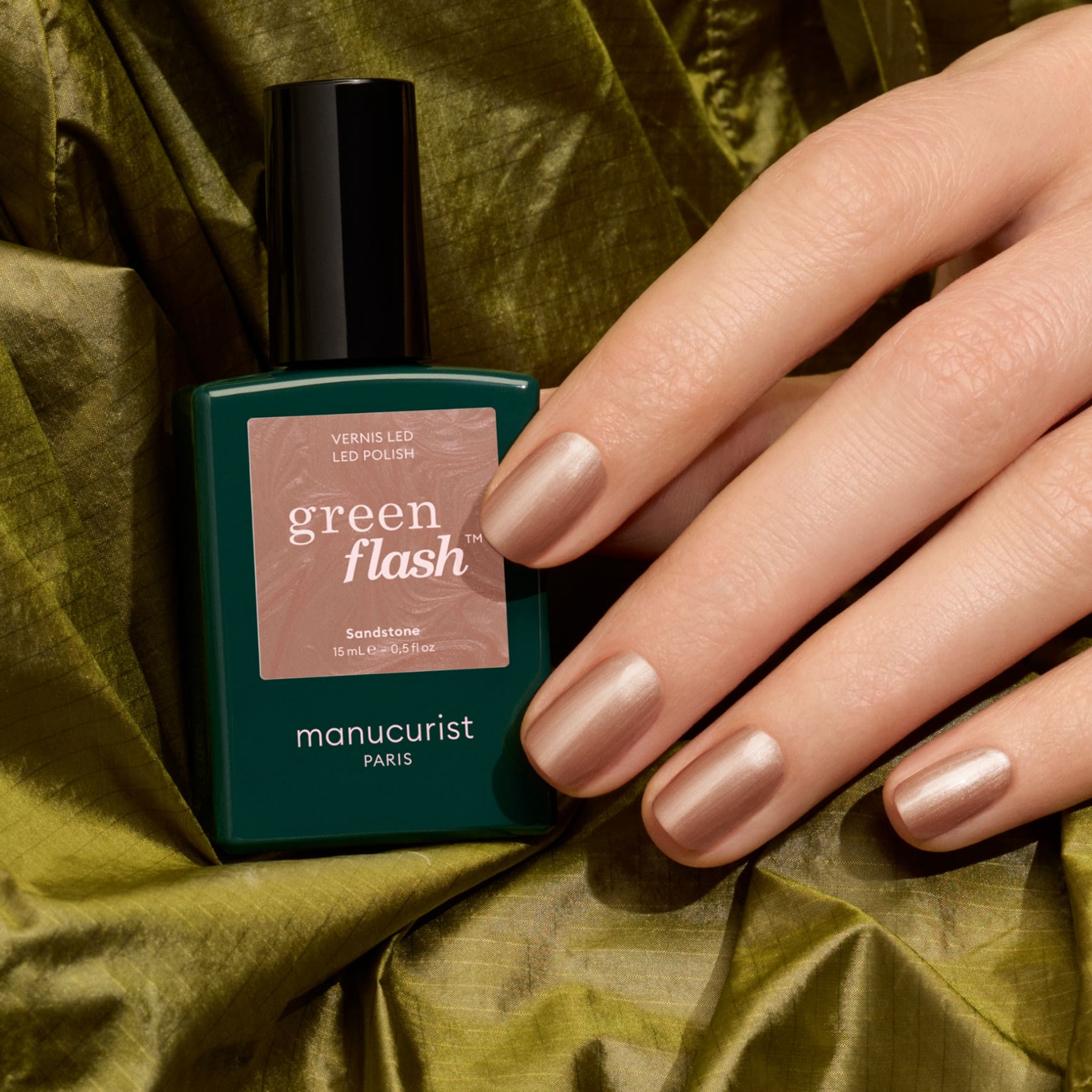 A hand with short, metallic beige nails holds a bottle of Sandstone gel polish by Manucurist, placed on shiny green fabric. The subtle sheen evokes the look of a shimmering latte brown or pearly brown gel polish.