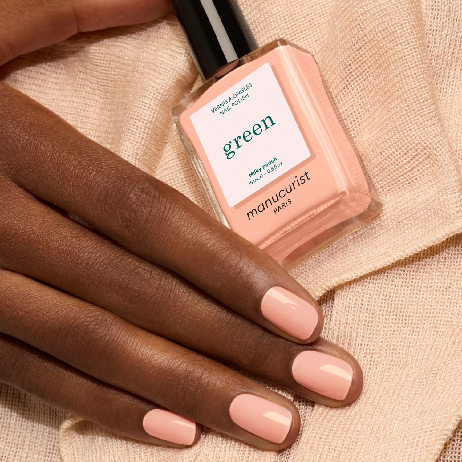 A hand with neatly manicured nails painted in Milky Peach rests on a peach fabric. Nearby is a bottle of Manucurist Milky Peach nail polish, its gentle shade perfectly complementing the soft elegance of the scene.