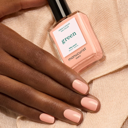 A hand with neatly manicured nails painted in Milky Peach rests on a peach fabric. Nearby is a bottle of Manucurist Milky Peach nail polish, its gentle shade perfectly complementing the soft elegance of the scene.