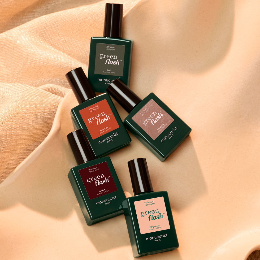 Five bottles from Manucurist’s Rainbow Collections are arranged diagonally on a soft beige fabric background, with warm lighting casting gentle shadows for a cozy effect. This curated set showcases vibrant manicure color palettes, perfect for pairing with your Green Flash LED lamp.