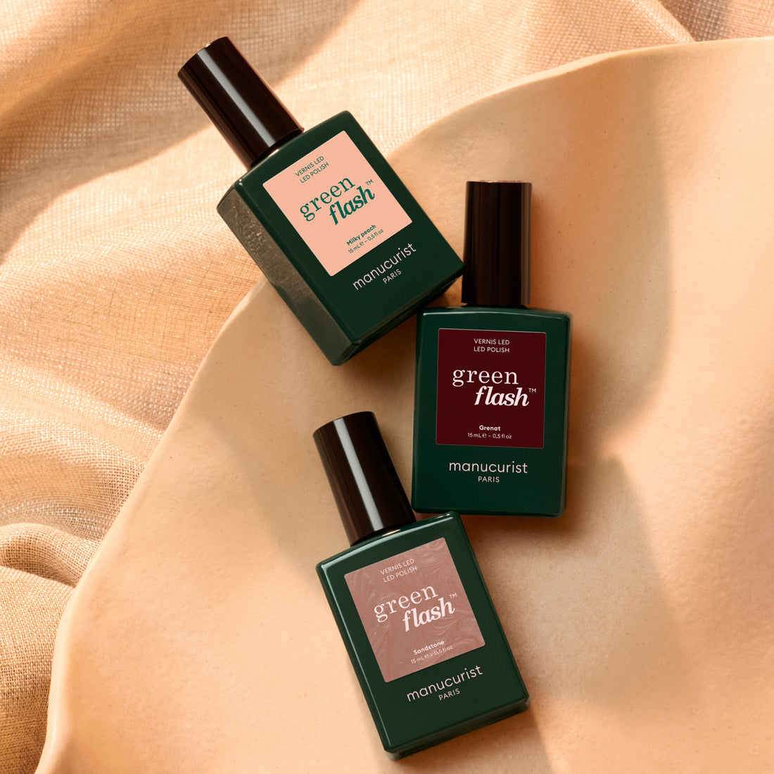 Three green bottles from the Manucurist Trio Collections are displayed on a beige surface, set against a soft beige fabric background. The labels highlight various curated shades and product names inspired by the latest nail trends.