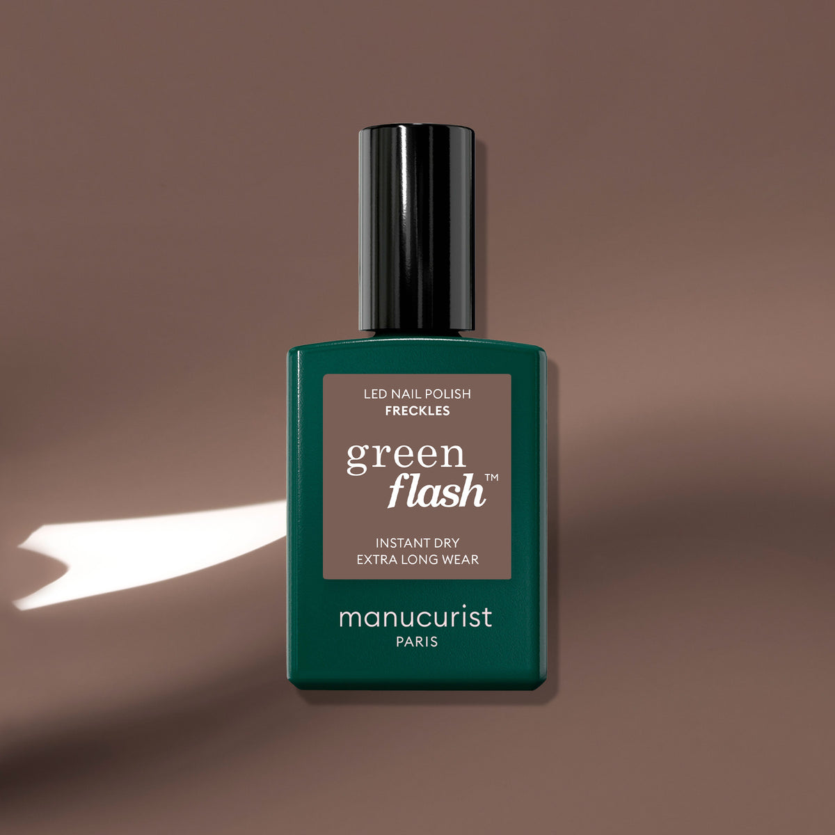 A green Manucurist Freckles nail polish bottle rests on a wood-toned brown surface with a streak of white light beside it.