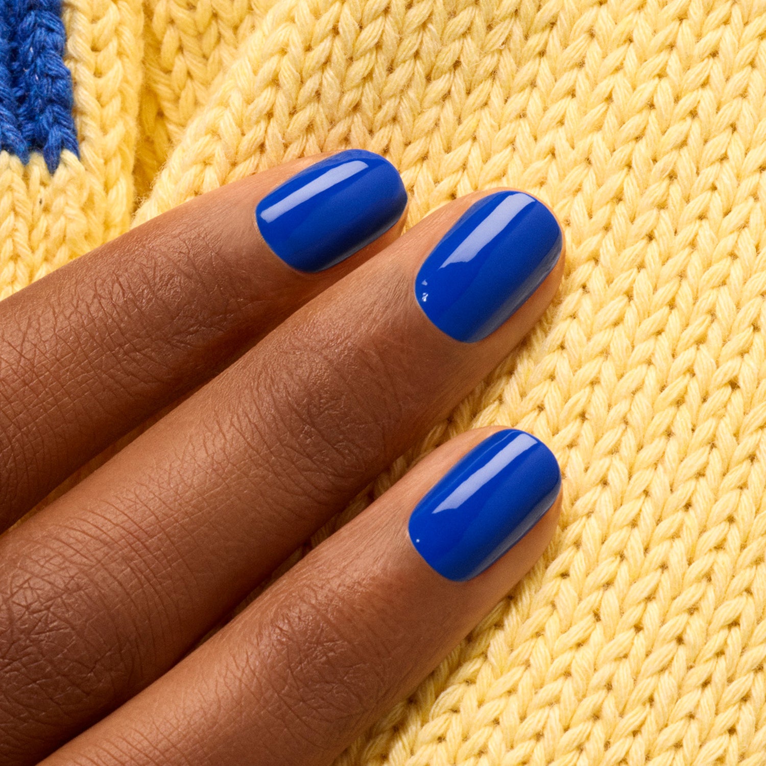 A hand with neatly manicured nails painted in Manucurist’s Ultramarine polish rests on yellow knitted fabric.