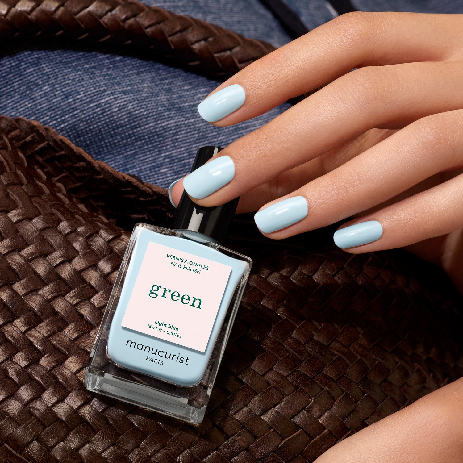 A hand with pastel blue nail polish holds a woven brown bag. Nearby, a bottle of Manucurist Light Blue nail polish rests on denim fabric.