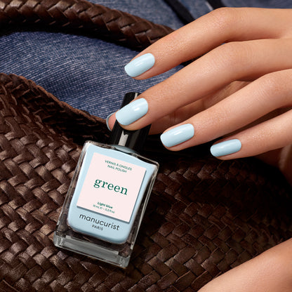 A hand with pastel blue nail polish holds a woven brown bag. Nearby, a bottle of Manucurist Light Blue nail polish rests on denim fabric.