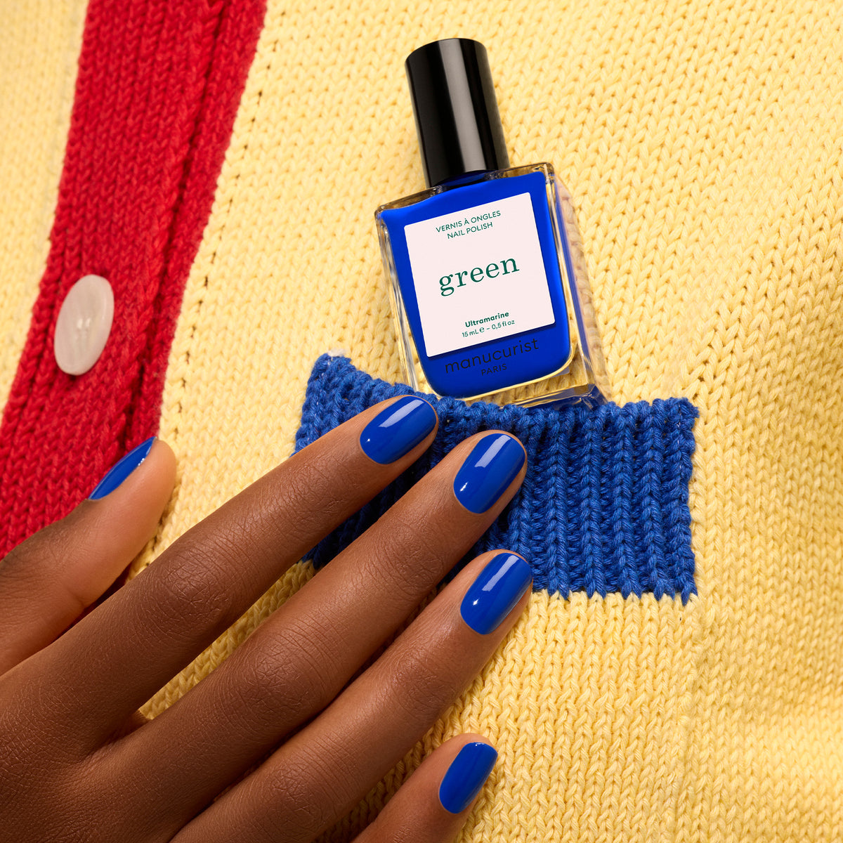 A hand with Ultramarine ocean blue nail polish holds a matching Manucurist bottle from the Green™ range, tucked into the pocket of a yellow and blue knit sweater with a red stripe and white button.