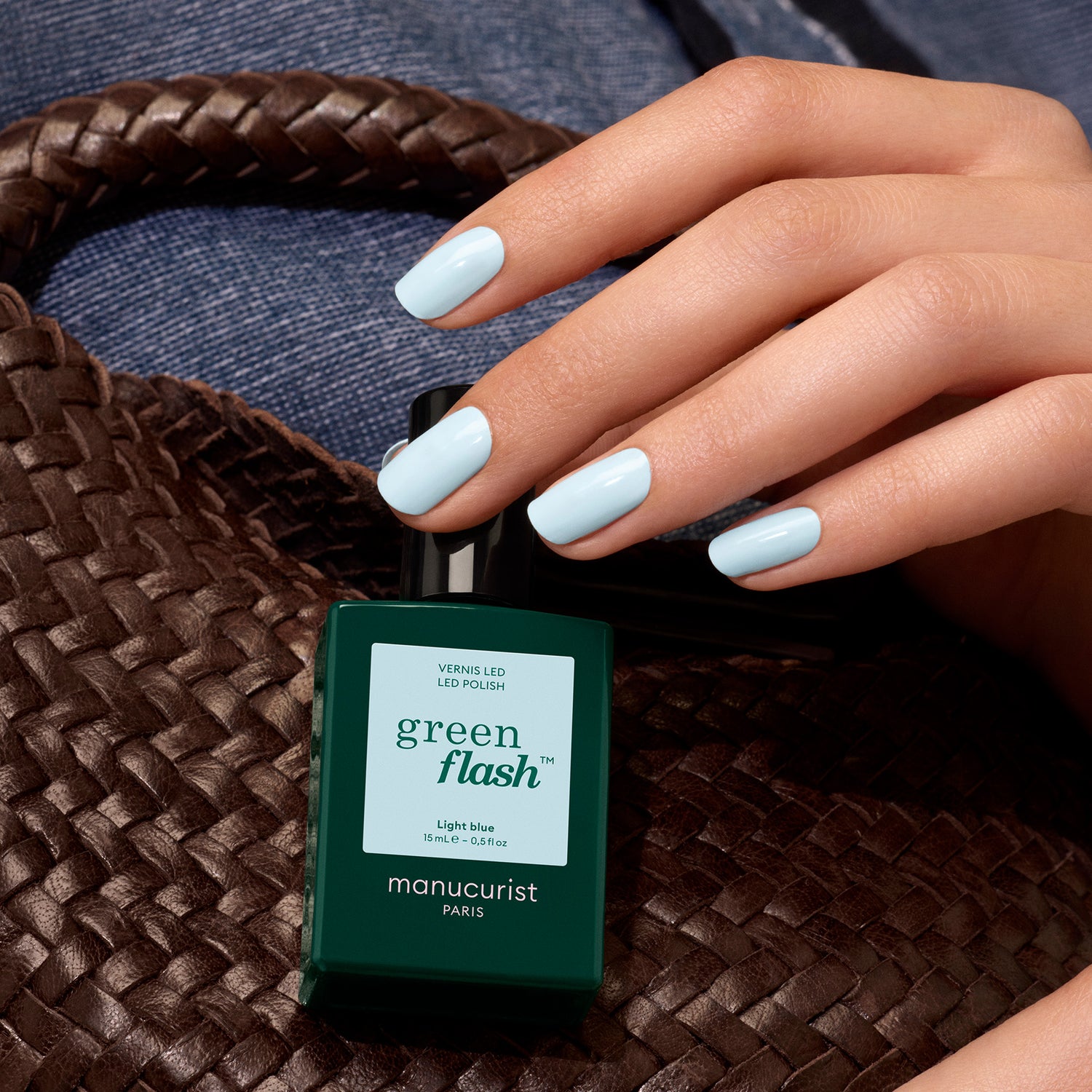 A hand with pastel blue nails holds a bottle of Light Blue gel polish by Manucurist, resting on a woven brown bag against blue denim fabric.
