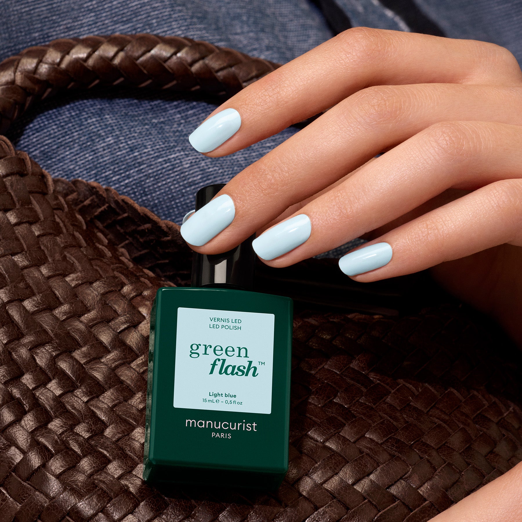 A hand with pastel blue nails holds a bottle of Light Blue gel polish by Manucurist, resting on a woven brown bag against blue denim fabric.