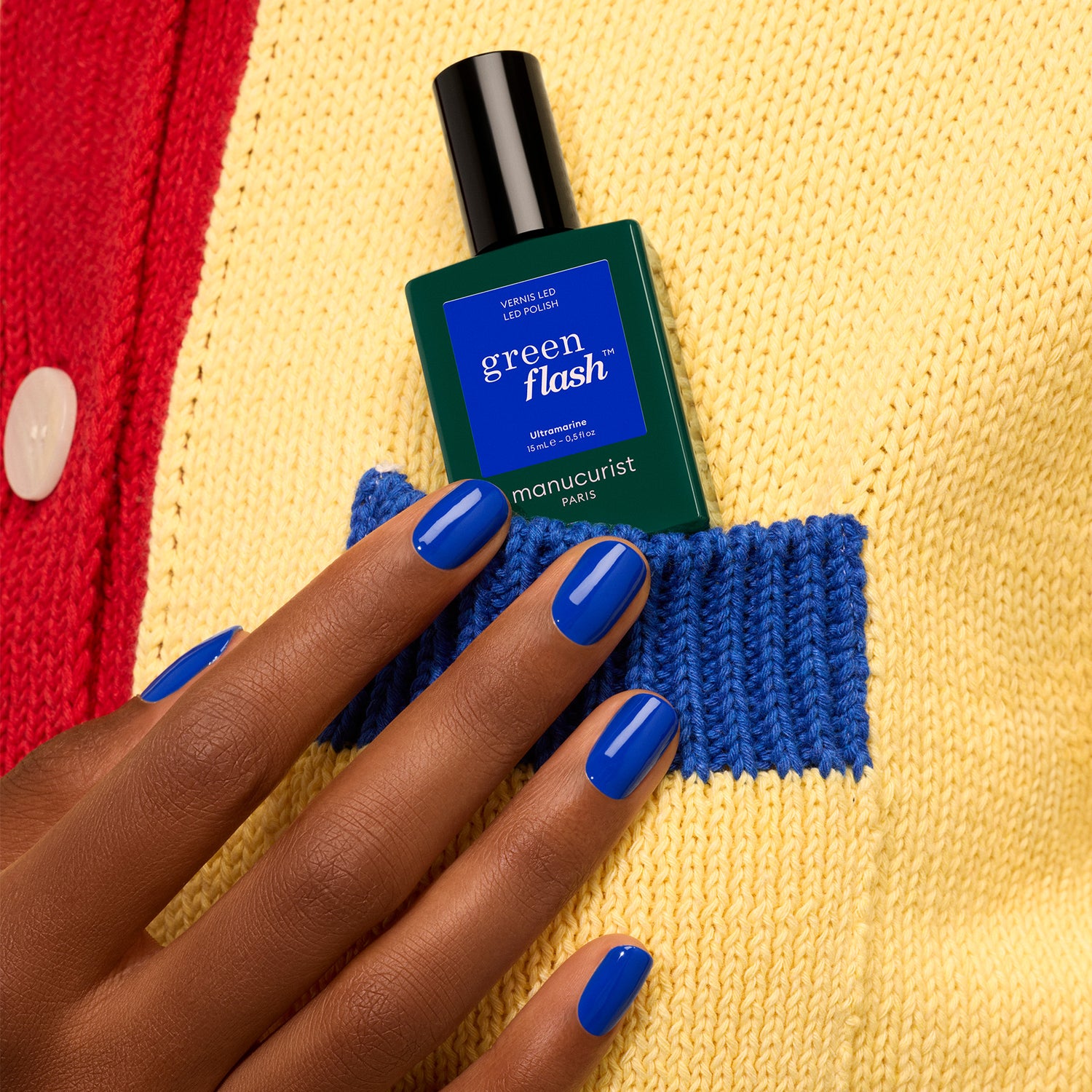 A hand with glossy electric blue gel polish (Ultramarine by Manucurist) holds a green bottle of Green Flash nail polish near the blue pocket of a yellow and red knitted sweater.