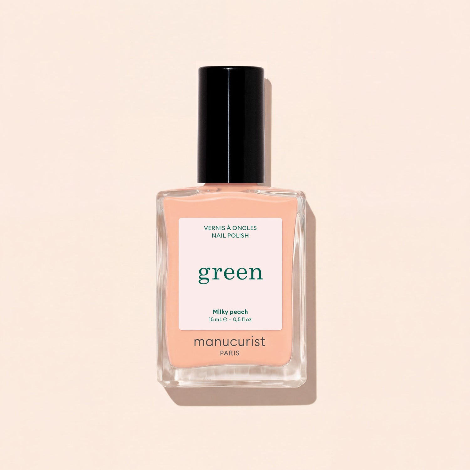 A bottle of Manucurist Milky Peach nail polish from the Green collection—a soft orange beige shade—featuring a black cap and “green” label, is displayed against a light beige background.