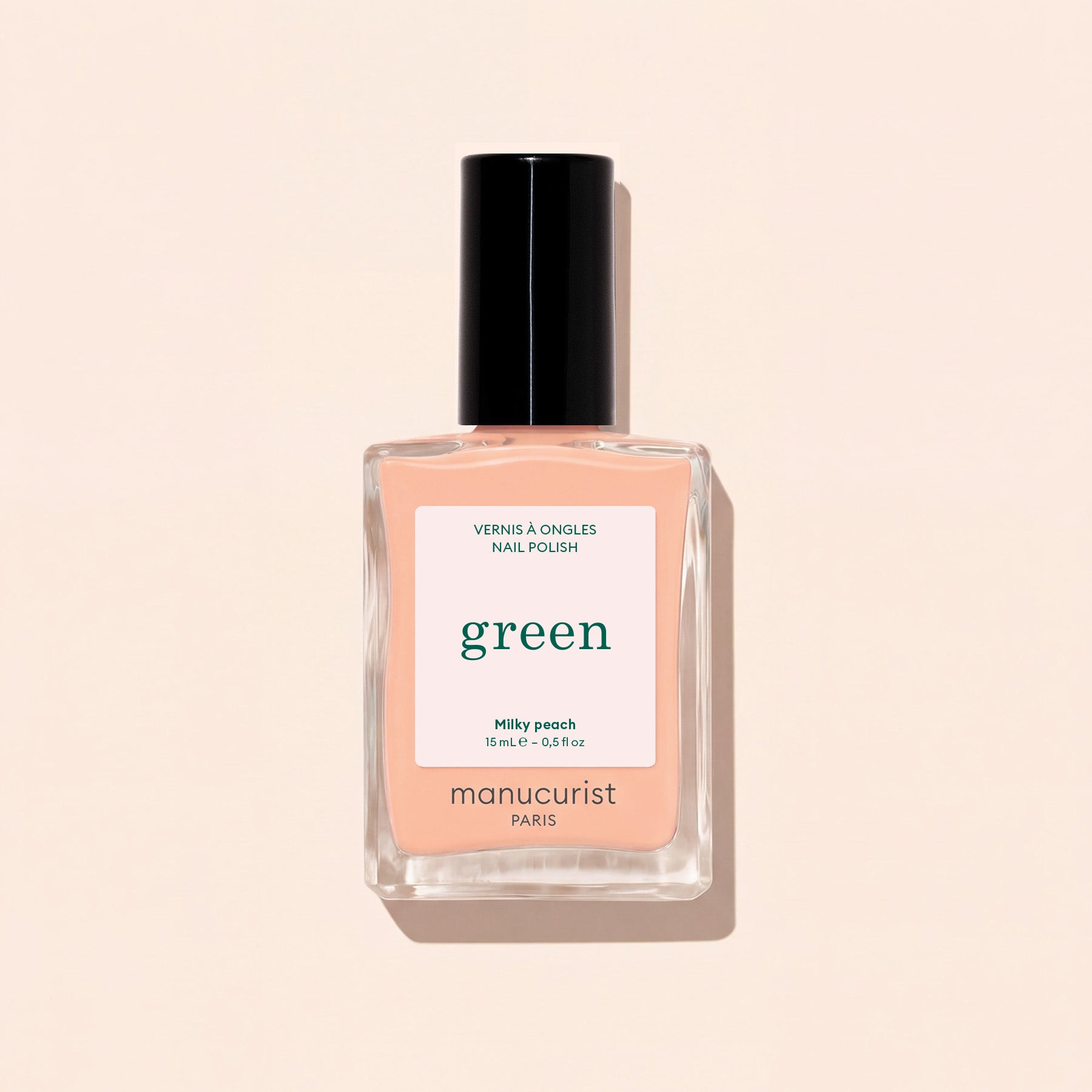 A bottle of Manucurist Milky Peach nail polish from the Green collection—a soft orange beige shade—featuring a black cap and “green” label, is displayed against a light beige background.