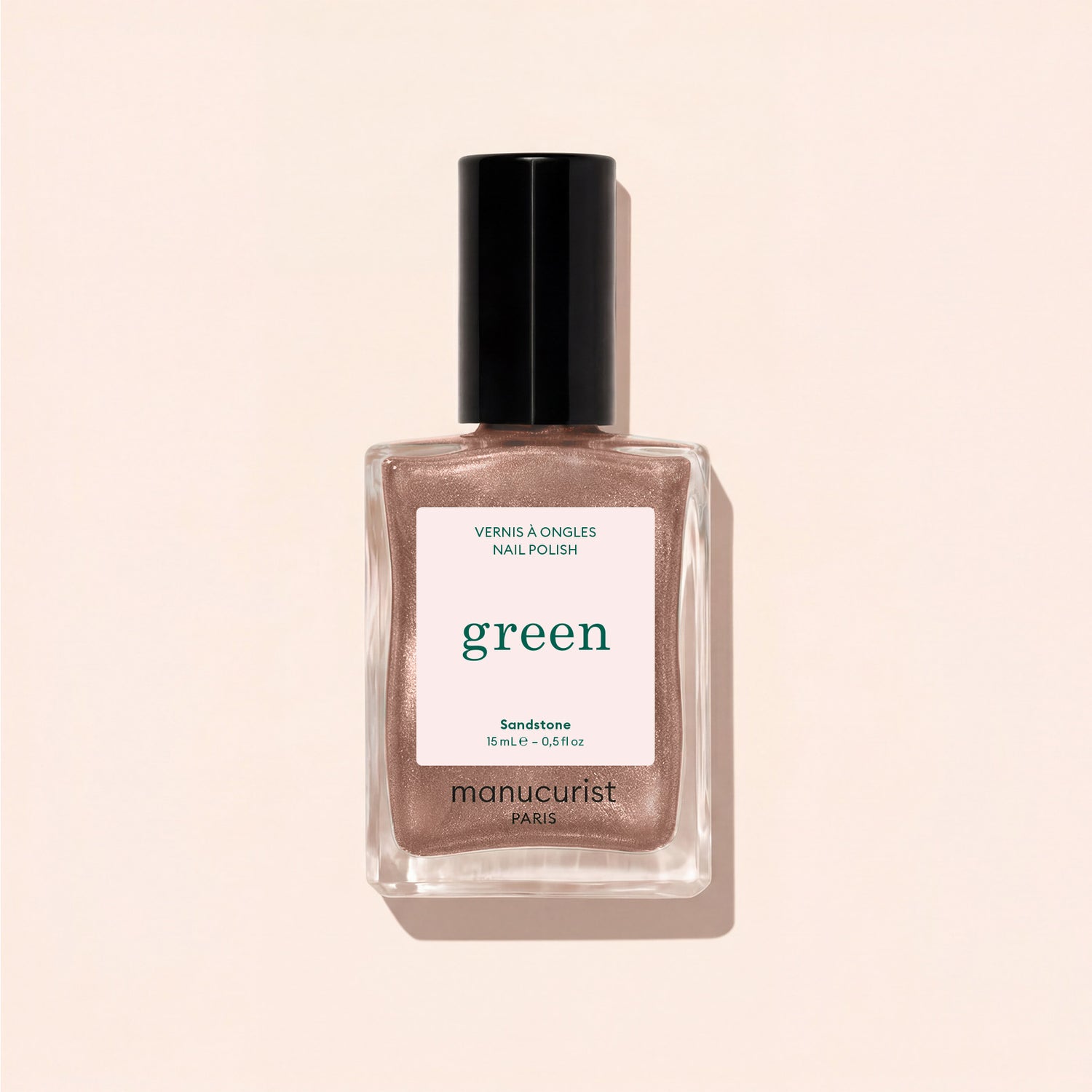 A bottle of Manucurist nail polish in the shade Sandstone is displayed on a light beige surface, with its shadow cast beside it.