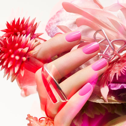 A close-up of a hand wearing bold silver rings, with Bloom by Manucurist—a neon pink polish with blue undertones—from the Green™ range, surrounded by pink and red flowers for a vibrant, elegant look.