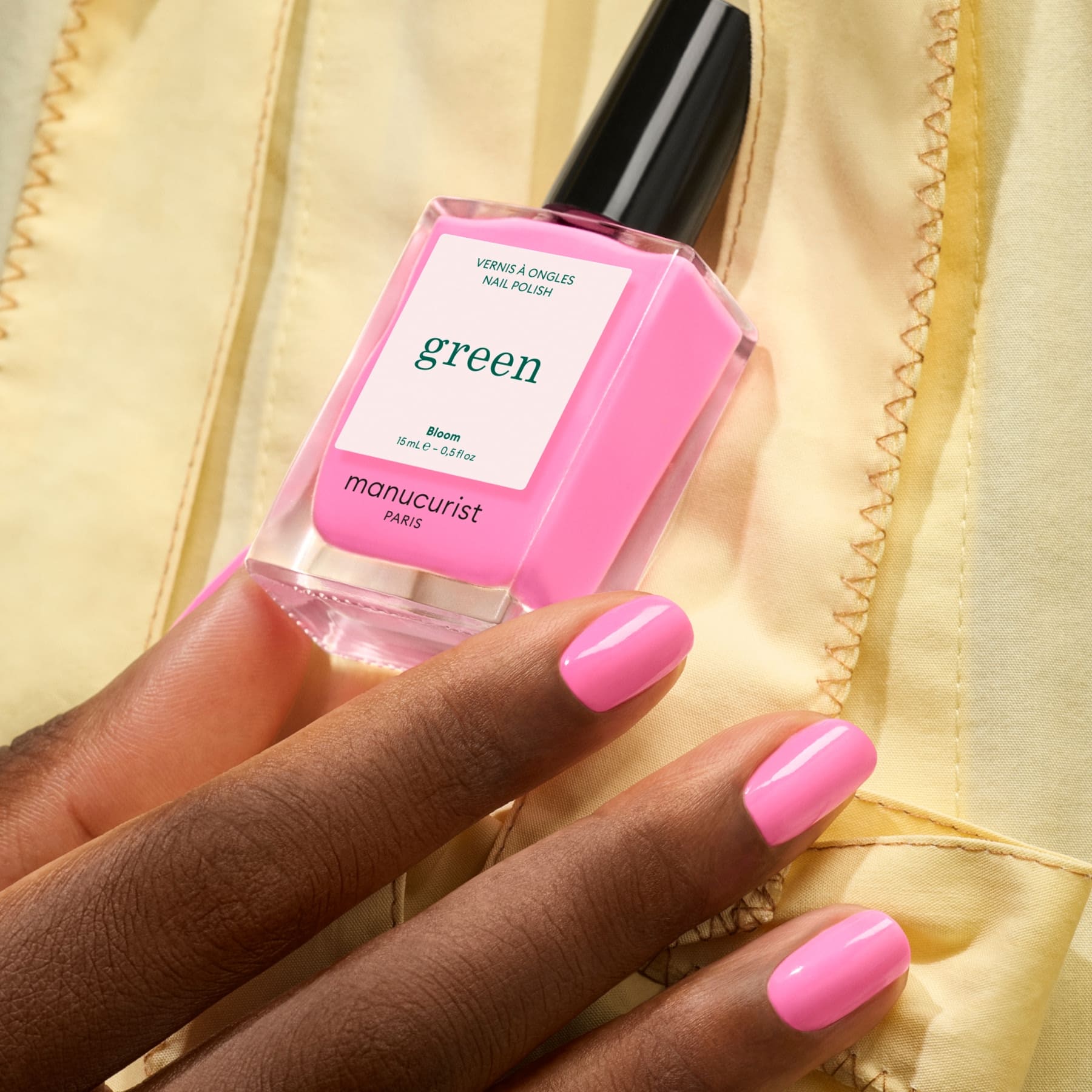 A hand with smooth, dark skin shows neatly manicured neon pink nails with blue undertones, holding a bottle of Bloom nail polish by Manucurist, against a pale yellow fabric background.