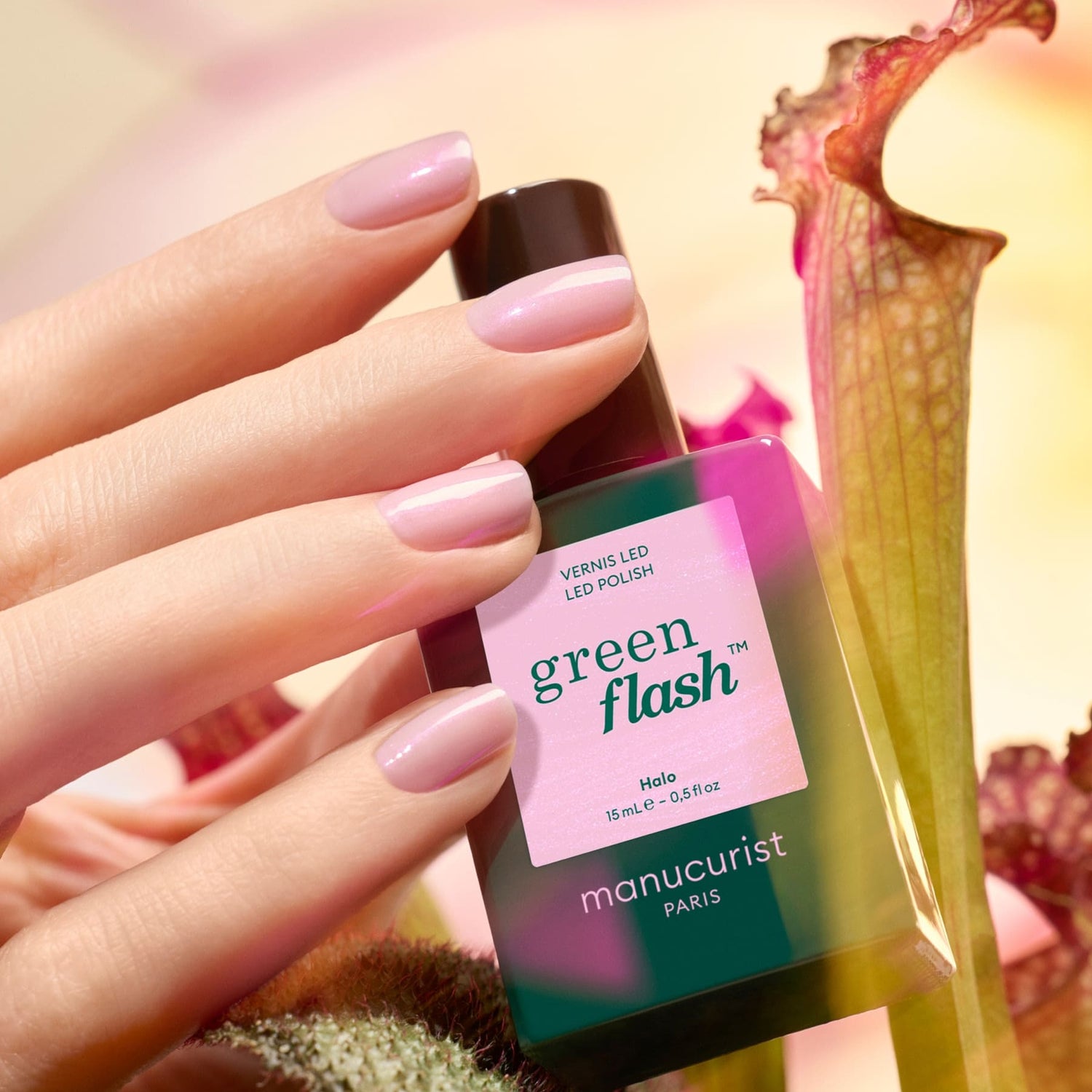 A hand with neatly manicured, light pink nails holds a bottle of Manucurist Halo polish. The background features softly blurred green and pink plants.