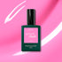 A bottle of Manucurist Bloom gel polish sits against a vibrant pink background with white streaks. The dark green bottle and pink label highlight the chic, magnetic intensity of this nail color.