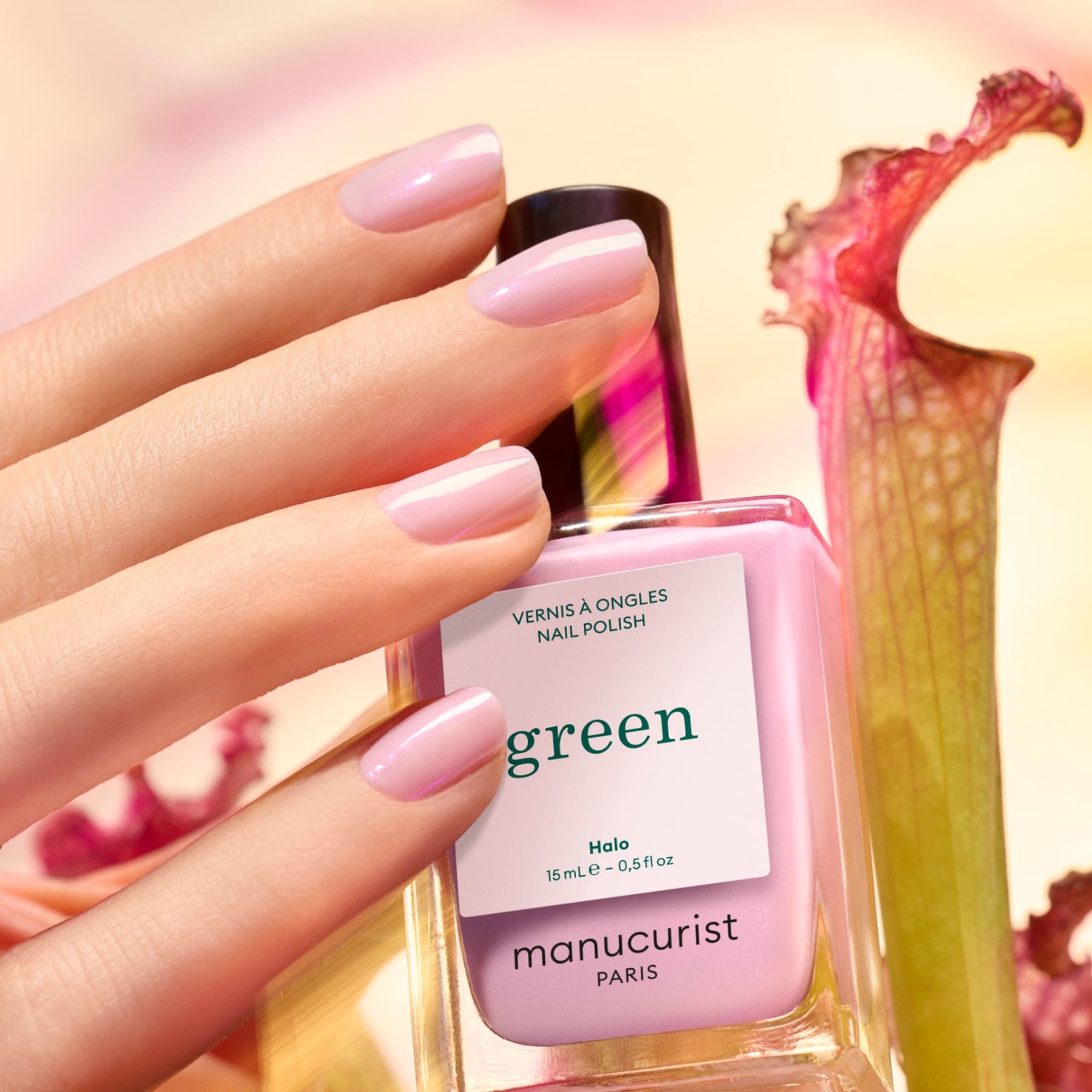 A hand with pastel pink nails holds a bottle of Manucurist Halo nail polish against a softly blurred background with light pink and green tones.