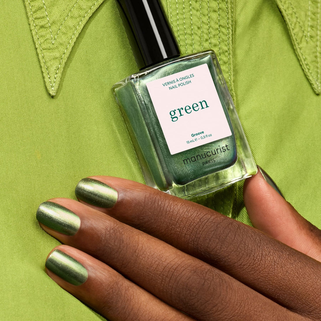 A hand with pearly green nail polish holds a matching Manucurist Groove bottle against a light green shirt, creating a stylish monochromatic look.