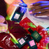 A hand with pastel-painted nails holds a white envelope labeled Manucurist above bottles of Rainbow Collections nail polish by Manucurist, vibrant anthurium flowers, and playful color palettes under colorful lighting.