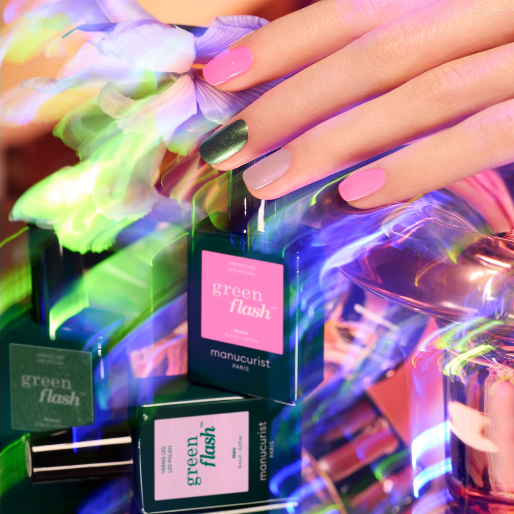 A hand with pink and metallic green nails, reflecting the latest trends, is shown above bottles of Manucurist Trio Collections nail polish, surrounded by colorful iridescent light and floral accents.