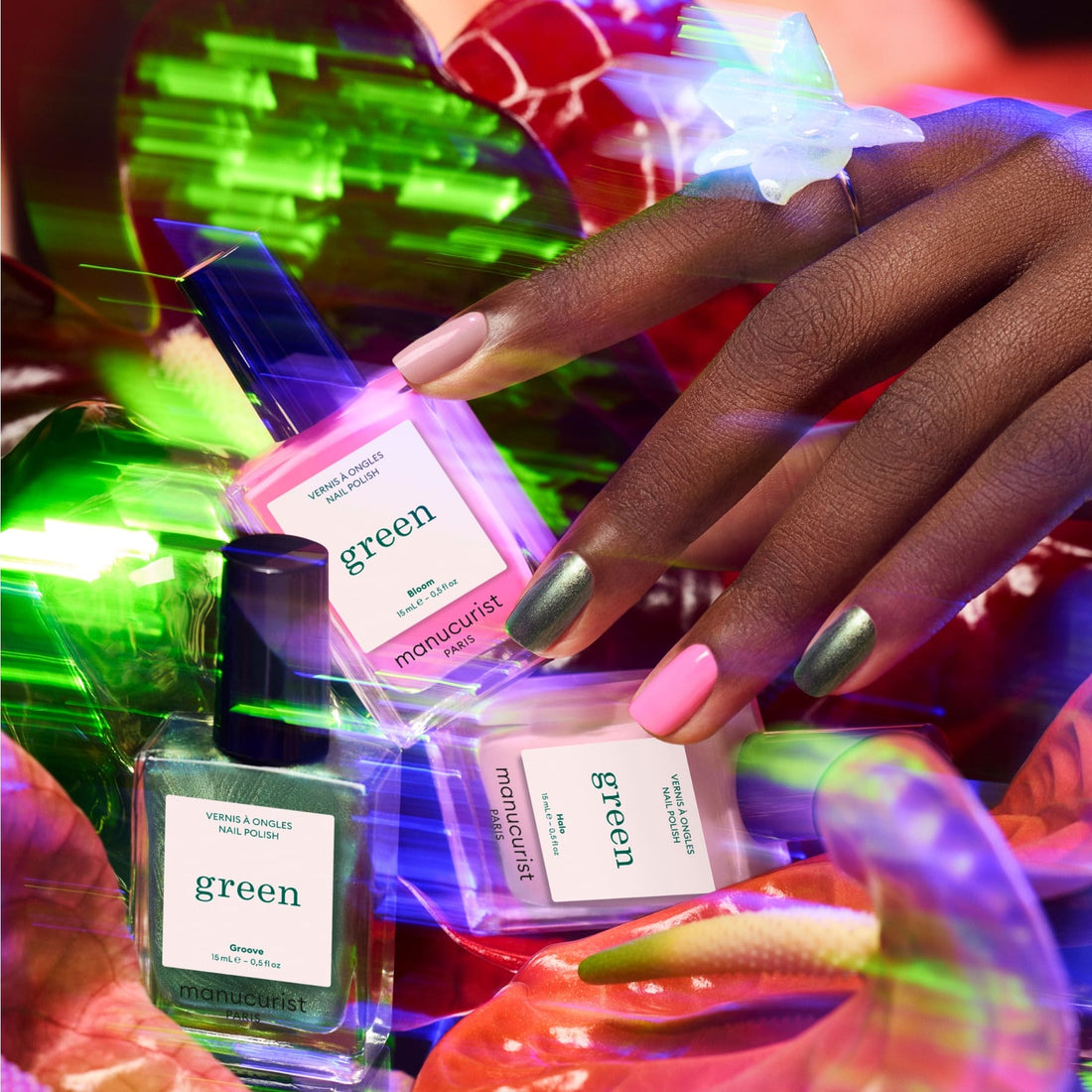 A hand with pink and metallic green nails holds a pink bottle from the Manucurist Trio Collections. Two more bottles are nearby on a vibrant surface featuring a large flower and butterfly ring, showcasing trendy nail looks.