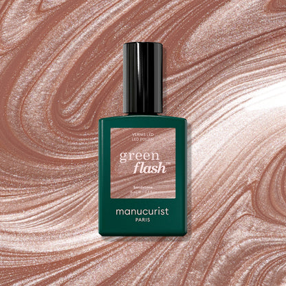 A bottle of Sandstone LED polish by Manucurist is displayed in front of a swirling, shimmering latte brown and beige background that matches the polish color.