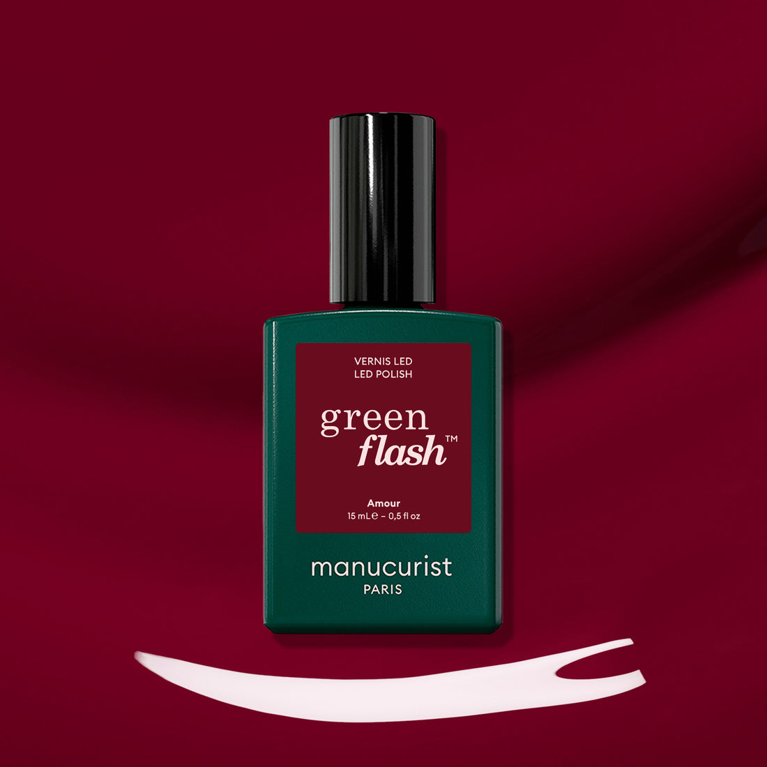 A bottle of Manucurist Amour gel polish rests on a deep burgundy background, with a swipe of luminous burgundy nail color displayed beneath it.
