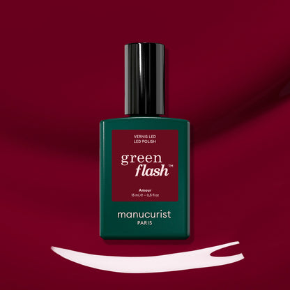 A bottle of Manucurist Amour gel polish rests on a deep burgundy background, with a swipe of luminous burgundy nail color displayed beneath it.