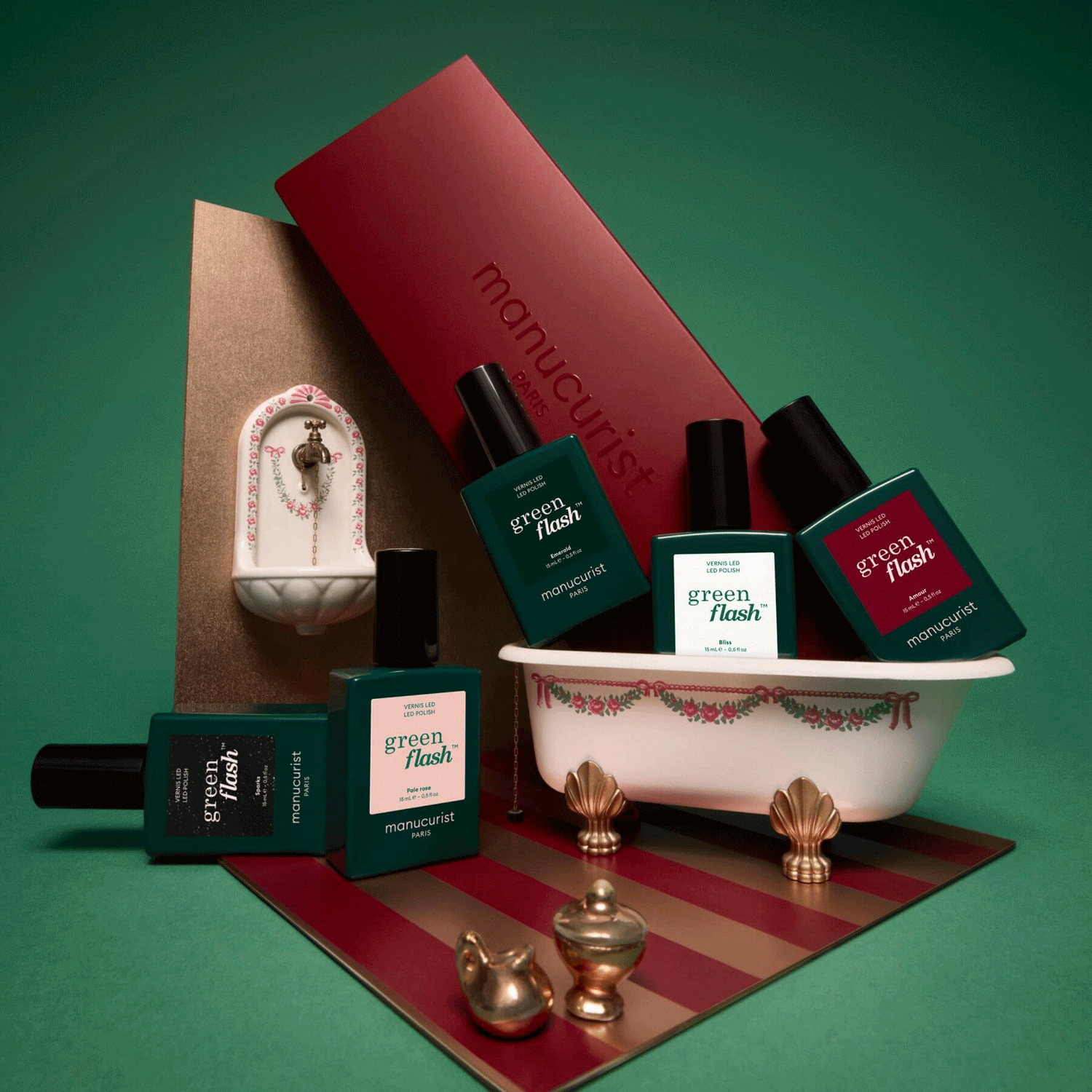 Bottles of Manucurist Customisable Rainbow Green Flash gel polish are arranged around a tiny decorative bathtub, faucet, and gold objects, set against a green background with a red Manucurist box.