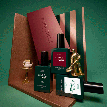 Three green bottles labeled Customisable Trio Green Flash by Manucurist are arranged with a gold figure, a small tea set, and striped and maroon props on a green background—perfect inspiration to create your own trio with their acetone-free gel polish remover for a complete manicure experience.